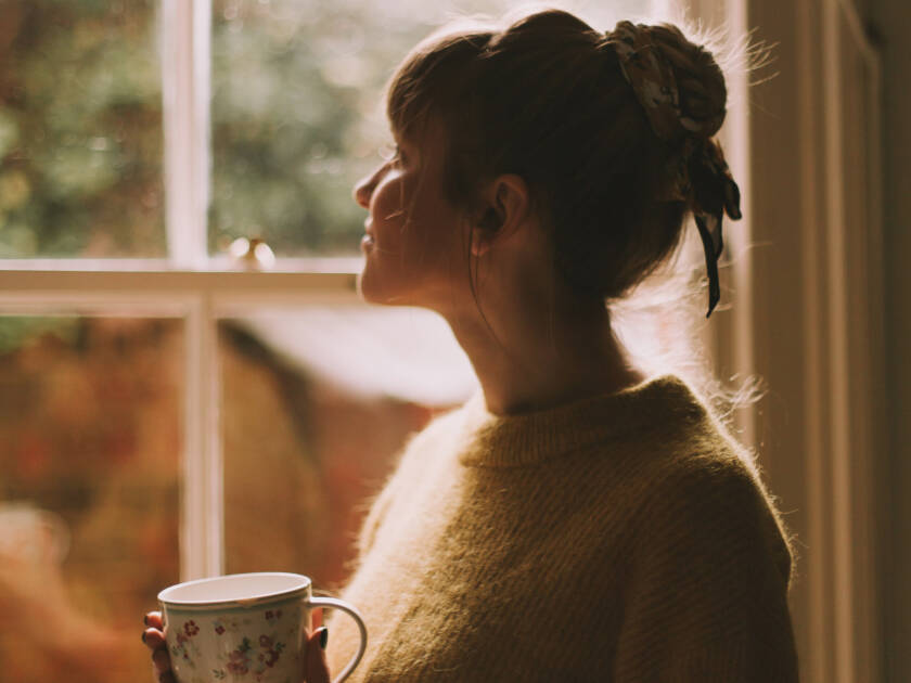 A woman is looking out a window and she is holding a mug in her hand.