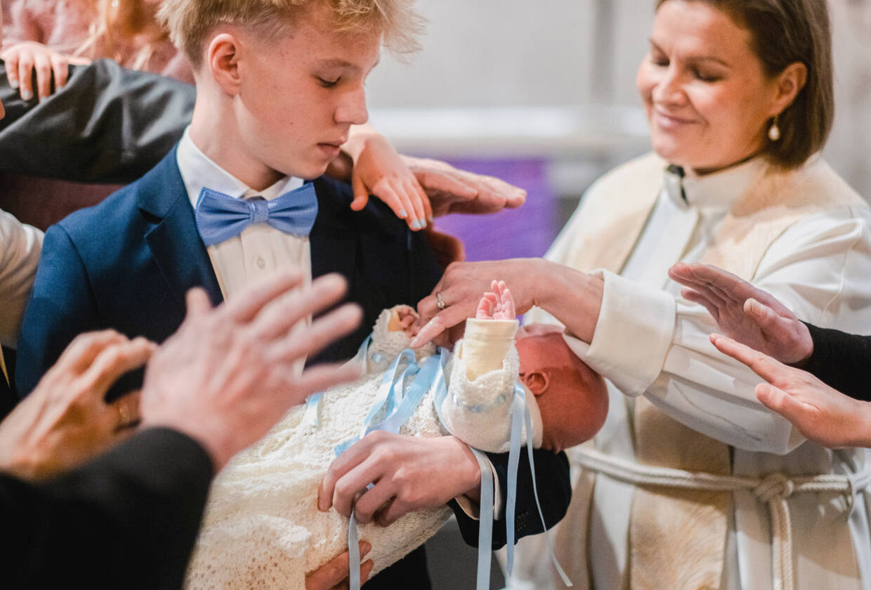 Kastettava vauva on nuoren pojan sylissä, ja pappi ja läheiset ovat nostaneet kätensä siunatakseen vauvaa.