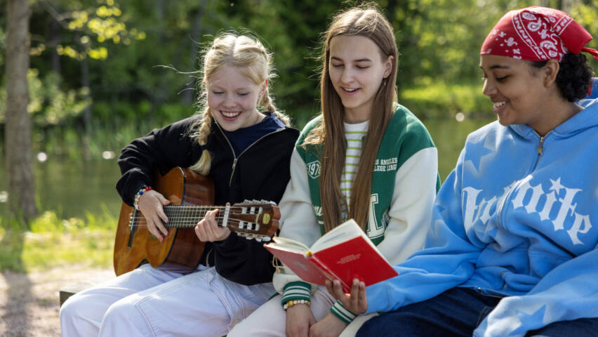 Tre flickor sitter ute och sjunger. En av dem spelar på en gitarr.